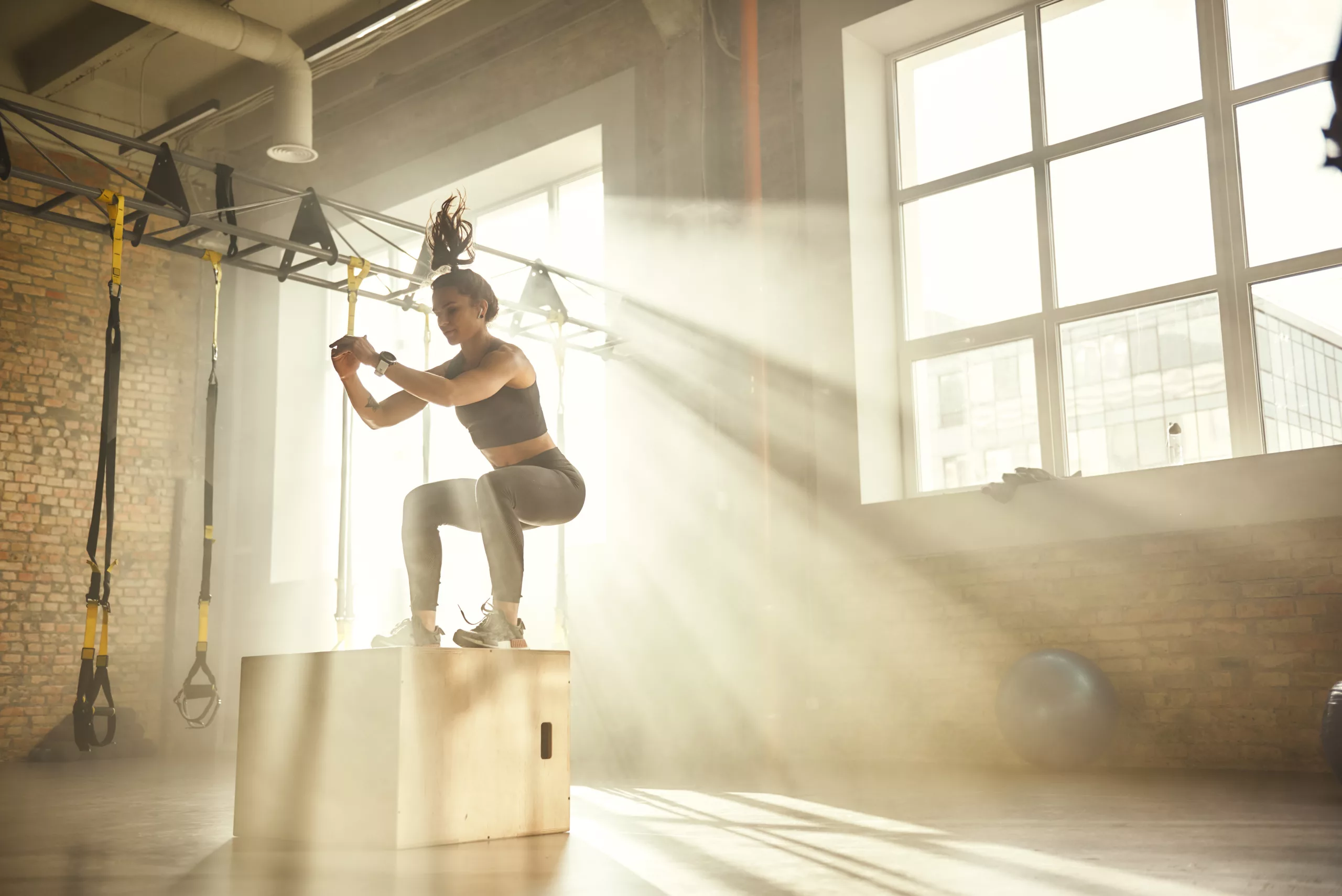Build Power & Athleticism with Plyometrics Side view of athletic woman in sportswear doing a box jump in gym with sun setting in window behind her