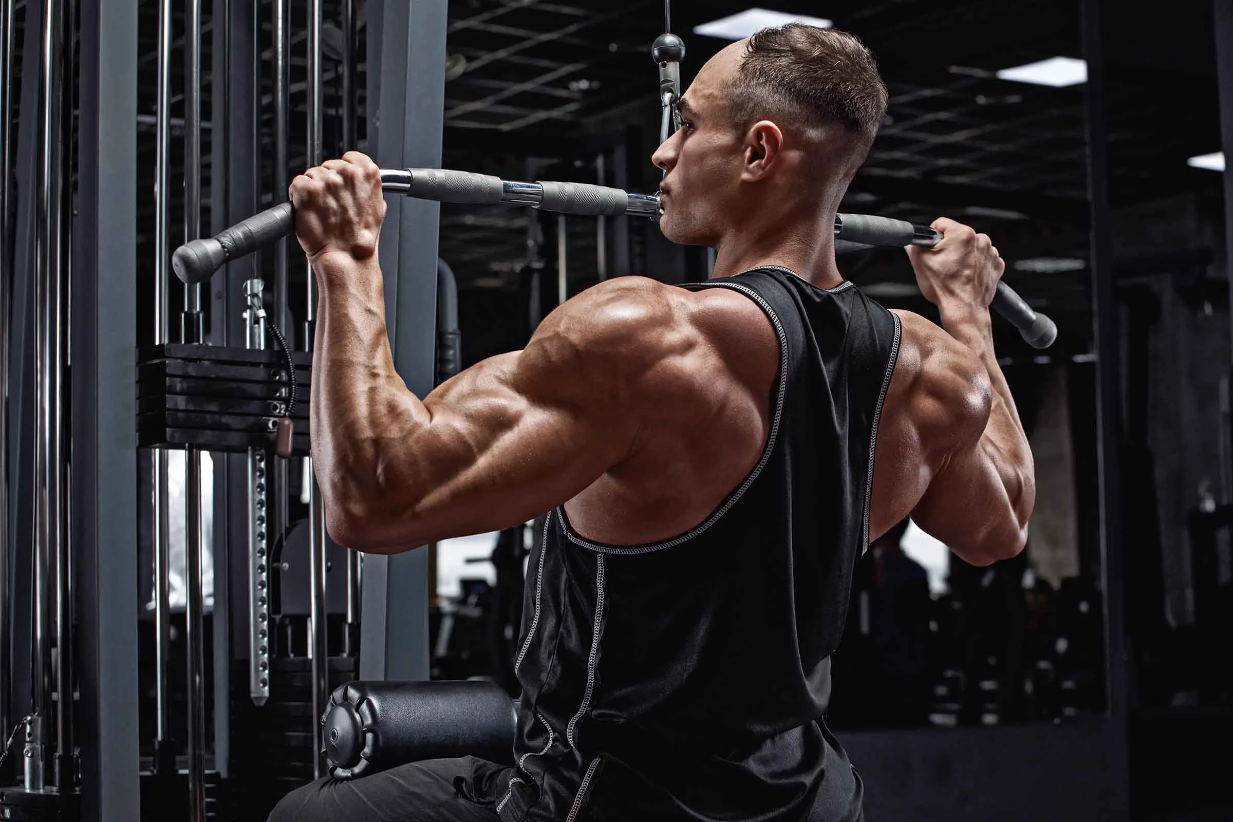 How & Why To Do More Lat Pulldowns Strong man in gym facing backward on lat pulldown machine doing lat workout